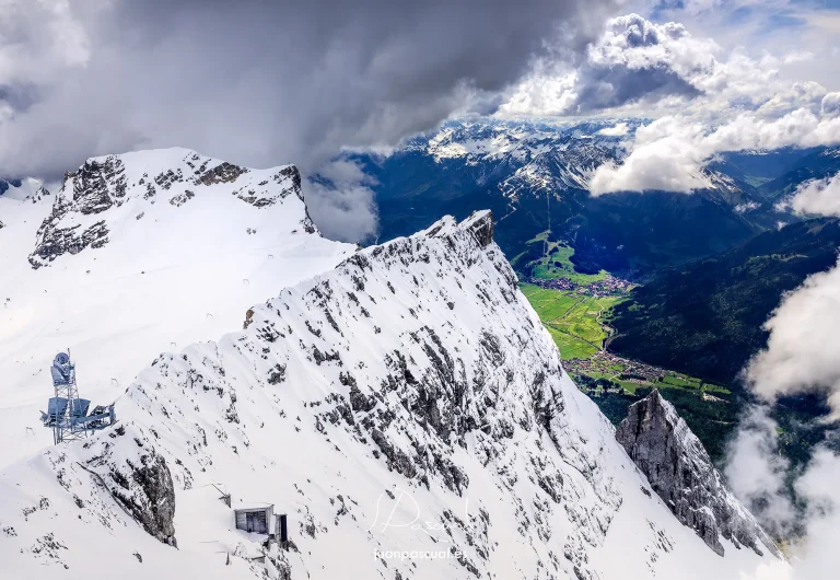 Impresionante panorama alpino desde las alturas del Tirol austríaco, donde las crestas nevadas dominan el primer plano mientras las nubes se deslizan entre los picos. Al fondo, el valle verde salpicado de pequeños pueblos contrasta con el blanco intenso de la montaña.