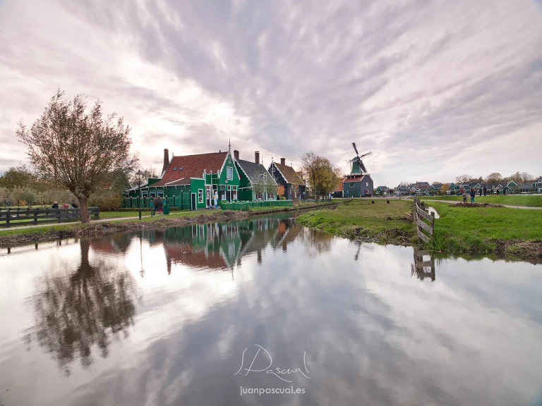 Paisaje tradicional de Zaanse Schans, en los Países Bajos, donde las casas de madera pintadas de verde y un molino de viento clásico reflejan su silueta en el tranquilo canal. Bajo un cielo dramático al atardecer, el conjunto crea una escena típica del campo holandés