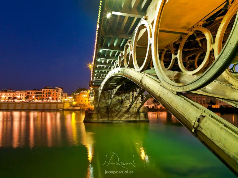 Fotografia realizada por el fotógrafo de arquitectura Juan Pascual: puente de Triana fotografiado de noche desde abajo, mostrando su estructura metálica con medallones circulares iluminada en verde y dorado, reflejada en las aguas del Guadalquivir, con el barrio de Triana al fondo. Sevilla, España.