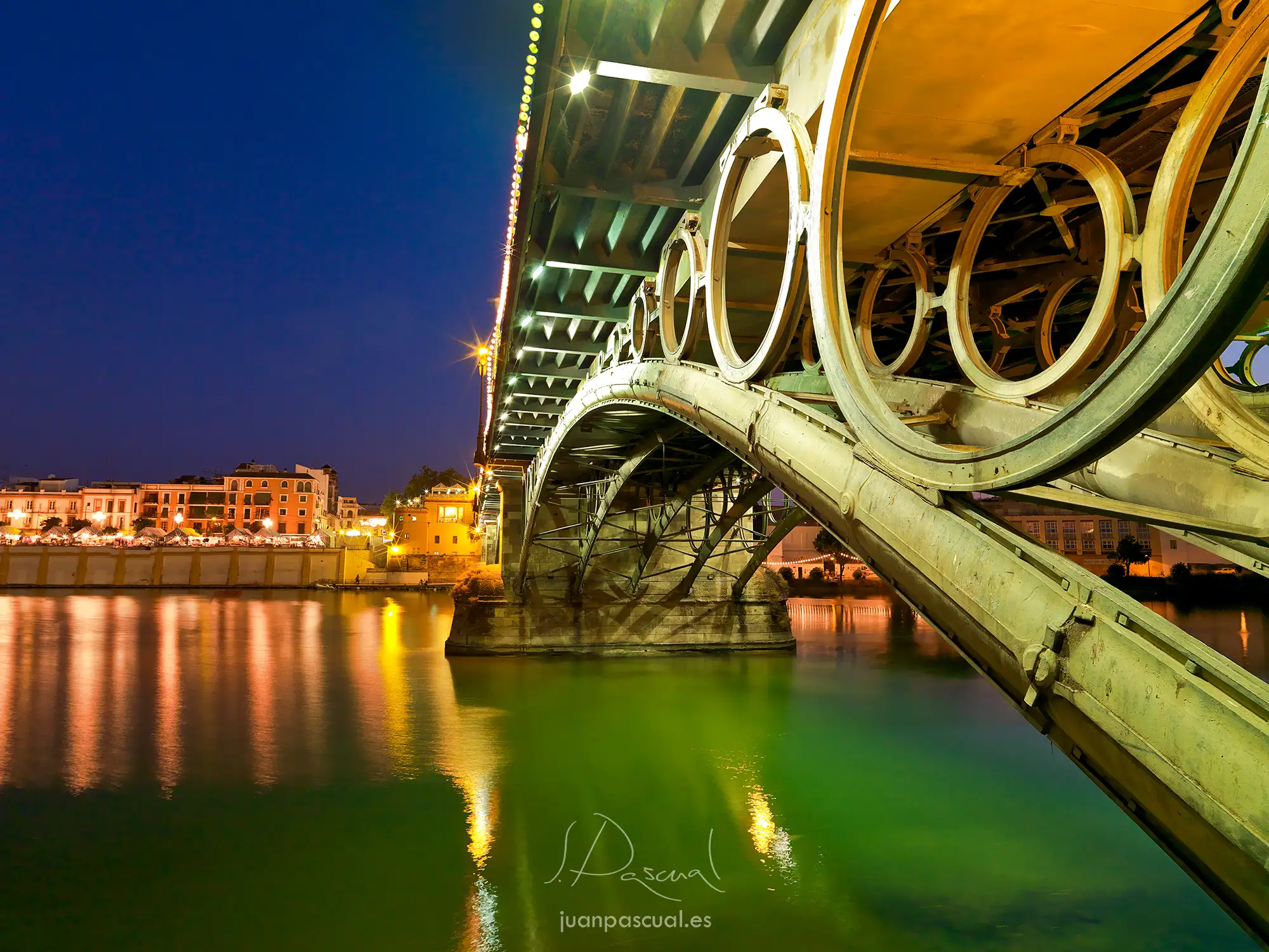 Fotografia realizada por el fotógrafo de arquitectura Juan Pascual: puente de Triana fotografiado de noche desde abajo, mostrando su estructura metálica con medallones circulares iluminada en verde y dorado, reflejada en las aguas del Guadalquivir, con el barrio de Triana al fondo. Sevilla, España.