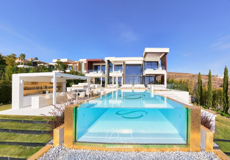 Vista frontal de una villa contemporánea en Marbella con gran piscina infinita y panel acristalado en primer plano. La terraza incluye zona de bar exterior y áreas de descanso, mientras la arquitectura moderna con amplios ventanales se abre al paisaje mediterráneo.