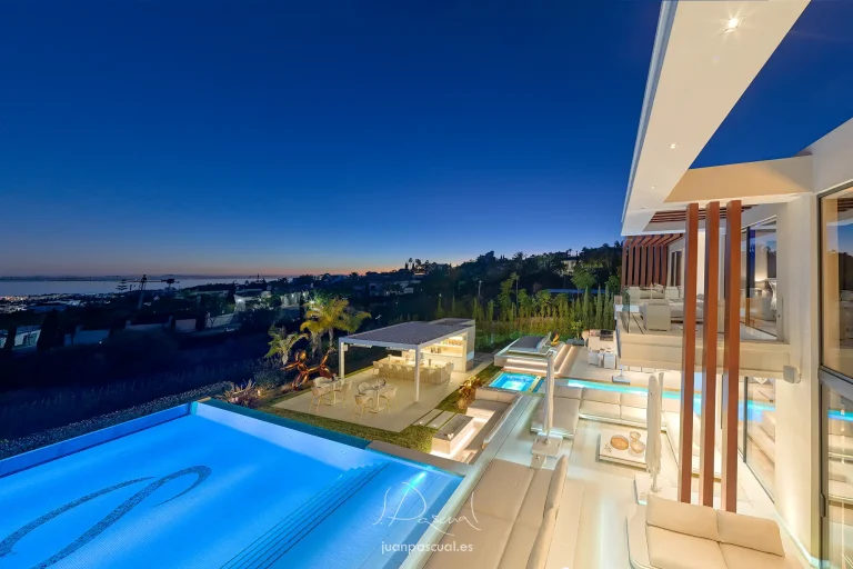 Fotografía de villa en Marbella, hecha por el fotógrafo inmobiliario Juan Pascual. Vista del exterior de la finca desde el dormitorio principal. Destacan la piscina, terraza de otro dormitorio, terraza de planta baja y zona de bar. El mar al fondo. Al anochecer.