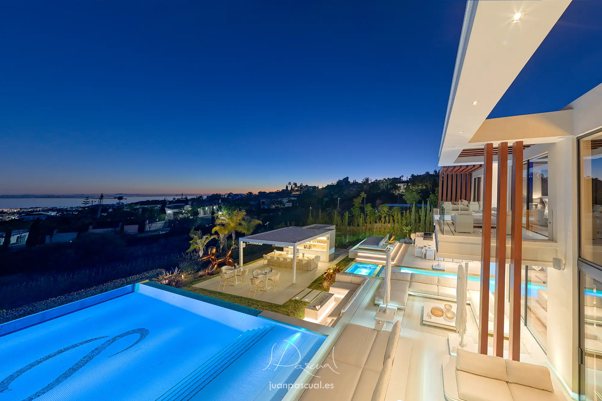 Fotografía de villa en Marbella, hecha por el fotógrafo inmobiliario Juan Pascual. Vista del exterior de la finca desde el dormitorio principal. Destacan la piscina, terraza de otro dormitorio, terraza de planta baja y zona de bar. El mar al fondo. Al anochecer.