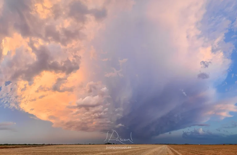 Tormenta espectacular sobre La Mancha, donde la inmensidad del cielo domina el paisaje. Una imponente nube convectiva despliega cortinas de lluvia y texturas dramáticas mientras la luz del atardecer tiñe las nubes de tonos rosados, dorados y azulados.