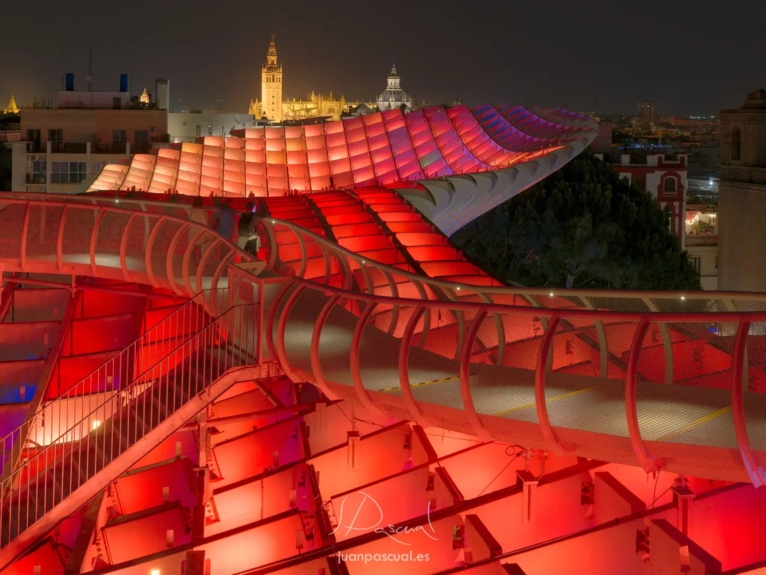 Fotografia nocturna de las Setas de Sevilla, hecha por el fotógrafo de turismo Juan Pascual. Iluminación en color rojo de la estructura superior con paseo de personas. La catedral al fondo.