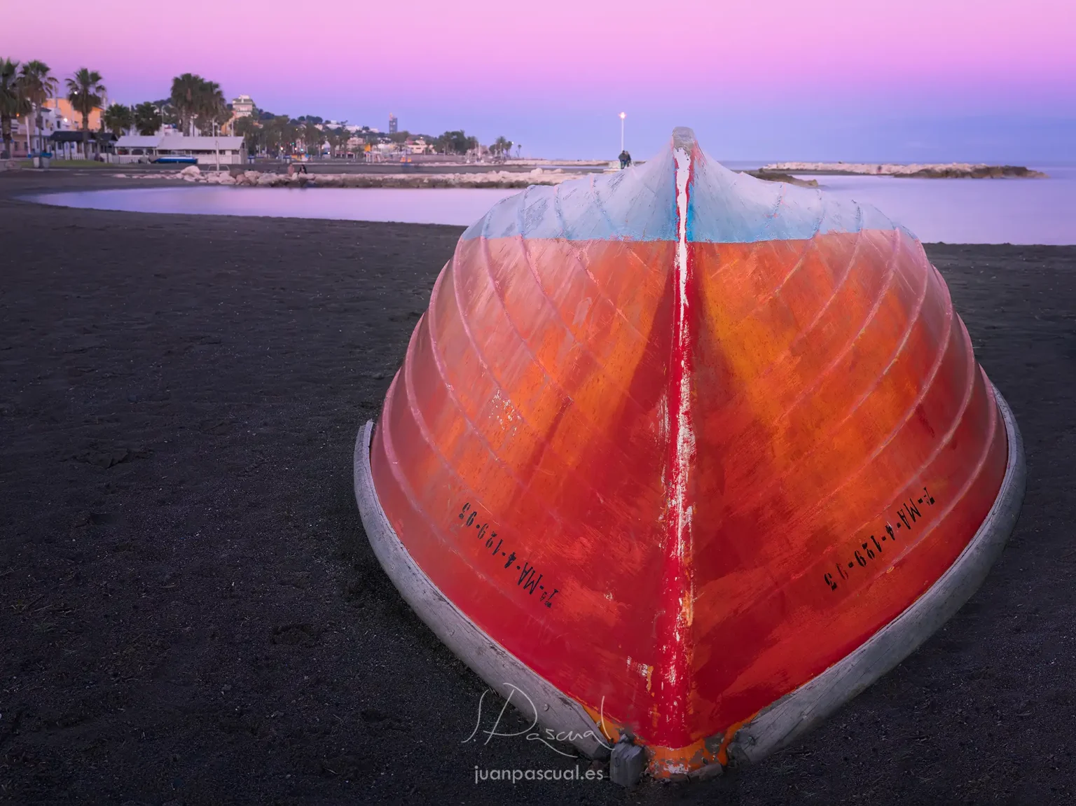 Barca roja de pescadores puesta del revés en la playa de Pedregalejo de Málaga, por el fotógrafo de turismo Juan Pascual. Hecha durante el ocaso de un día de primavera. El barrio de pescadores está al fondo.