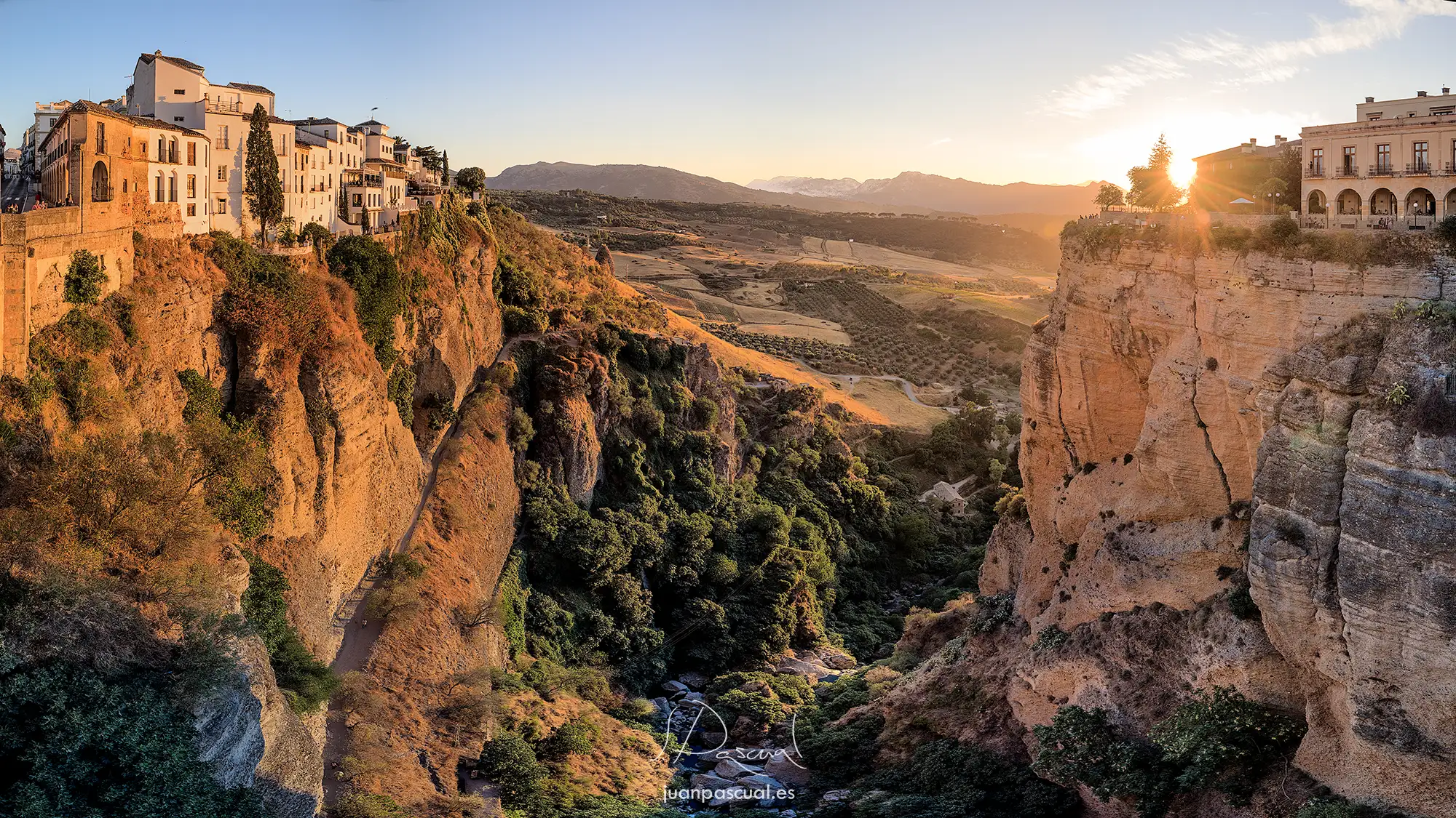 Fotografía realizada por el fotógrafo de turismo Juan Pascual: Fotografía de gran angular del Puente Nuevo de Ronda al atardecer. Se observa el imponente arco de piedra sobre el desfiladero del Tajo, con las casas blancas del pueblo colgadas al borde del precipicio y las montañas de la sierra al fondo bajo un cielo despejado.