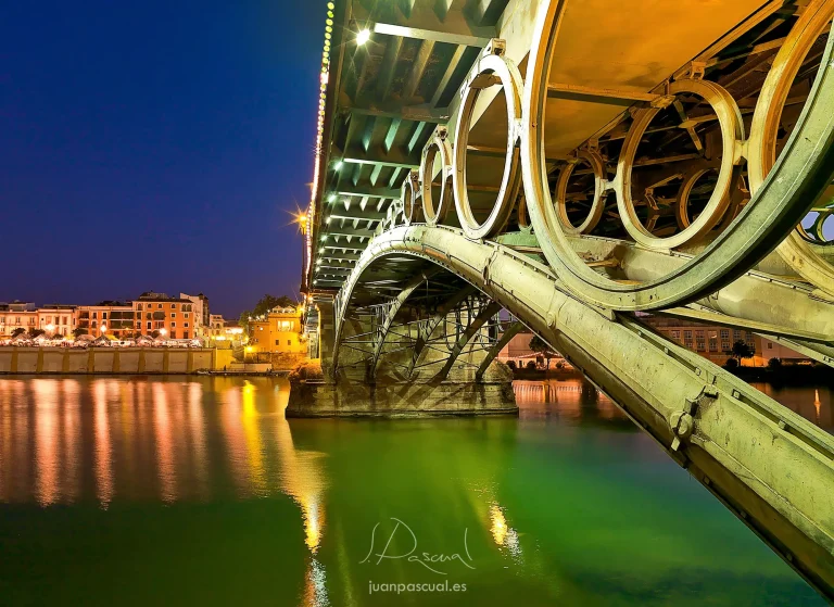 Vista nocturna del Puente de Triana (Puente de Isabel II) en Sevilla, fotografiado desde una perspectiva baja que resalta la estructura metálica y sus característicos aros decorativos. Las luces cálidas de la ciudad se reflejan en las aguas tranquilas del río Guadalquivir,
