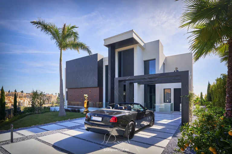 Entrada de una villa contemporánea en Marbella con fachada geométrica en blanco y negro, grandes ventanales y palmeras en el jardín. Reportaje hecho como fotógrafo de arquitectura. Un coche descapotable negro está estacionado en el camino de acceso, rodeado de paisajismo mediterráneo bajo un cielo azul luminoso.
