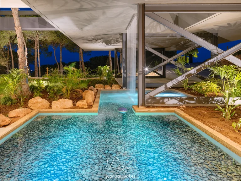 Zona exterior de una villa contemporánea en Sotogrande al anochecer, con láminas de agua decorativas bajo una estructura arquitectónica de acero y hormigón. La iluminación paisajística resalta plantas tropicales, rocas y árboles, creando un ambiente elegante y sereno.