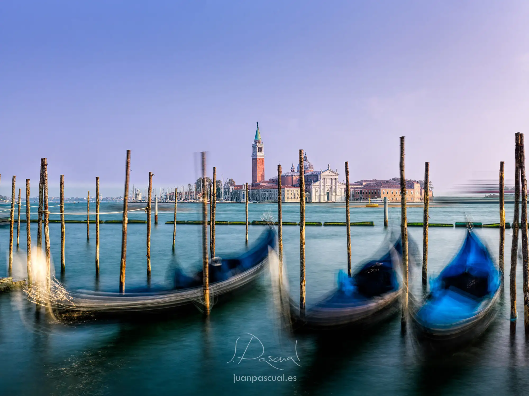 Tres góndolas están amarradas a postes de madera en aguas tranquilas, con un desenfoque de movimiento que muestra su desplazamiento. Al fondo, la iglesia de San Giorgio Maggiore y su campanario se ven a través de la laguna de Venecia, Italia.